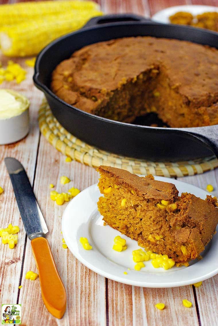 A piece of gluten free cornbread on a white plate with a cast iron skillet of cornbread, a knife and a bowl of butter.