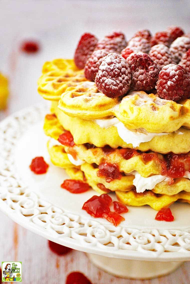 Closeup of a waffle cake with cream cheese, jam, and sugared raspberries on a white cake stand.