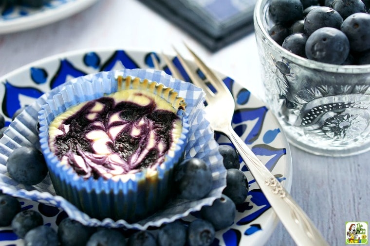 A plate with a mini blueberry cheesecake cupcake with blueberries and fork with a cup of blueberries.