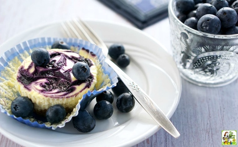 A small white plate with a mini cheesecake with a fork and glass cup of blueberries.