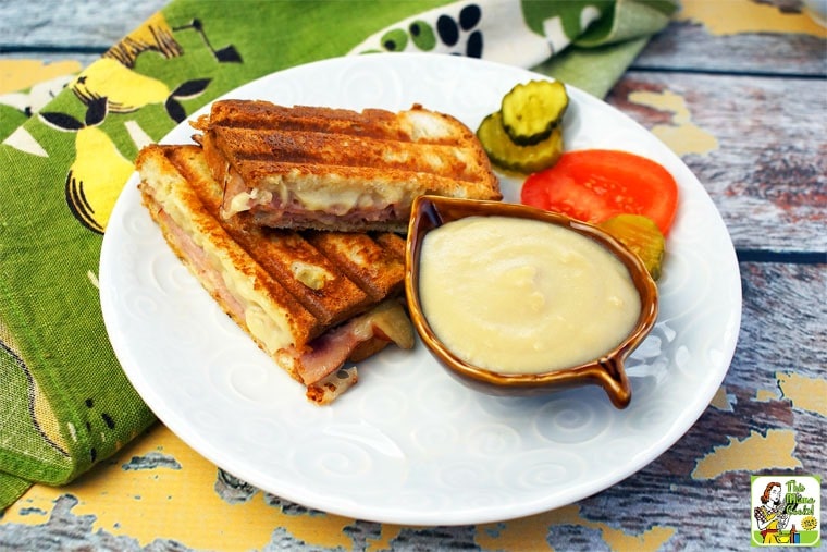 A white plate a grilled ham and cheese Croque Monsieur sandwich, a bowl of bechamel sauce, pickles and sliced tomatoes
