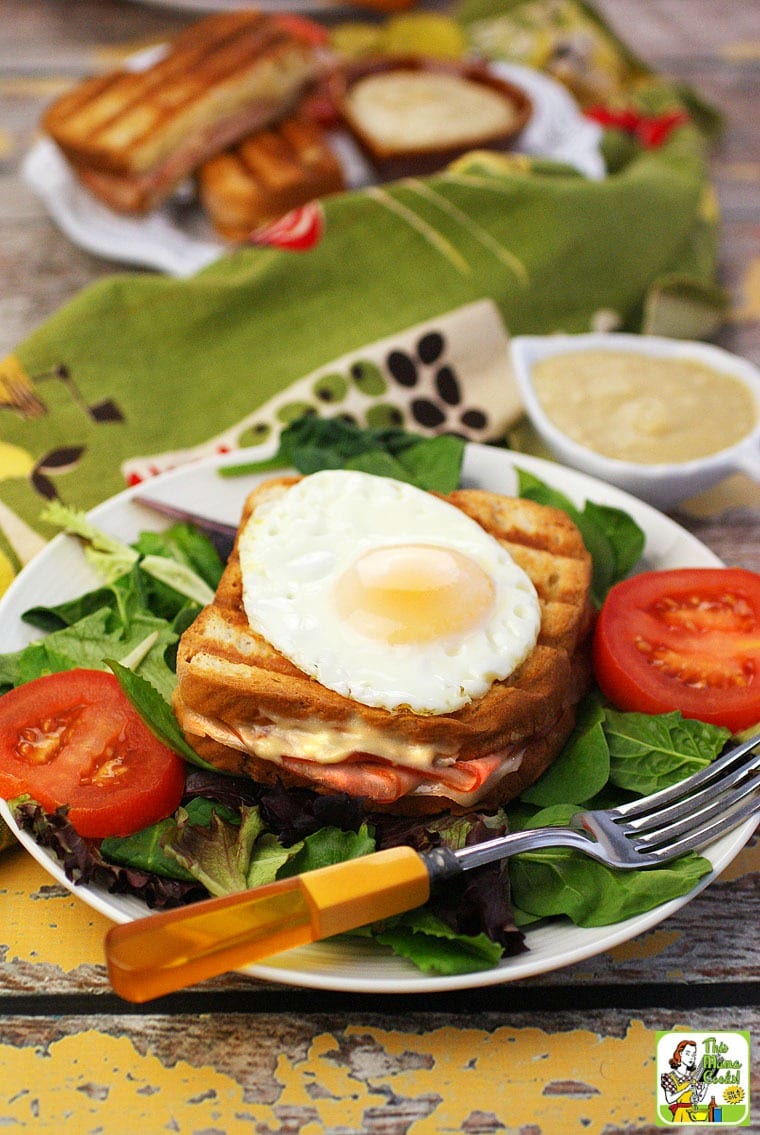 A plate with lettuce, a grilled Croque Monsieur sandwich with an egg, a bowl of bechamel sauce, and sliced tomatoes.