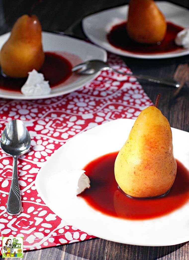 Slow Cooker Poached Pears in cherry sauce with a dollop of coconut whipped cream on white plates with spoons and napkins.