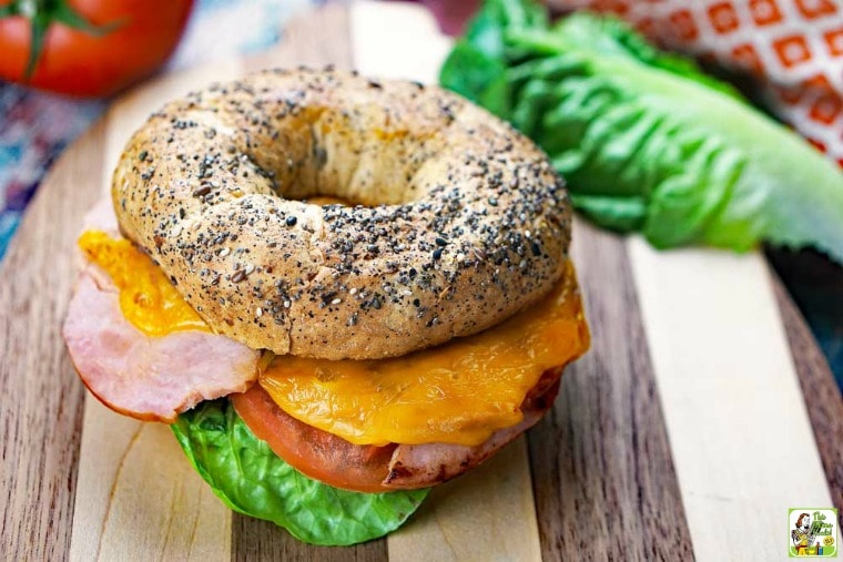 A breakfast bagel sandwich with layers of lettuce, tomatoes, ham, and cheese on a wooden cutting board.