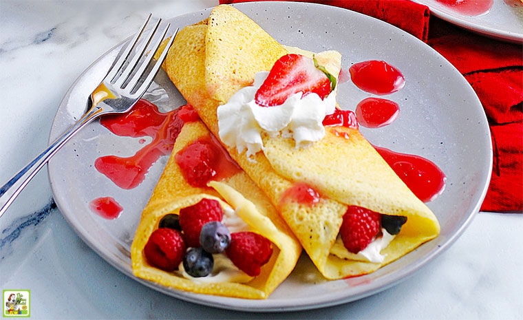 Two gluten free crepes filled with creme and berries covered in whipped cream and strawberry sauce on a grey plate with a fork.