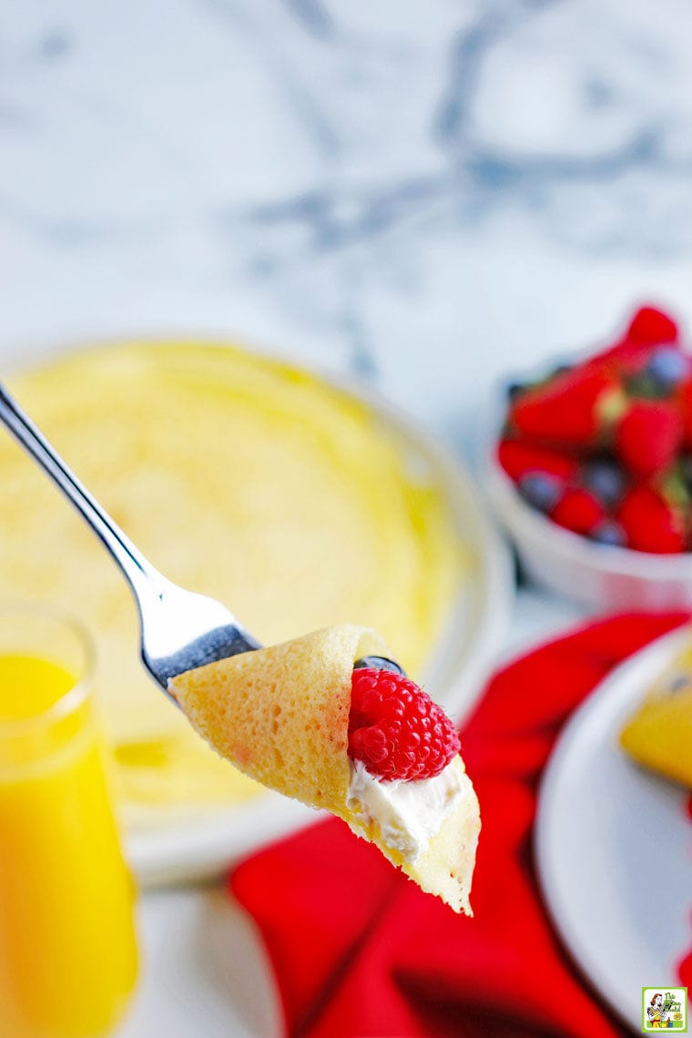 A forkful of cream and raspberry filled gluten free crepe with berries and crepes in the background.