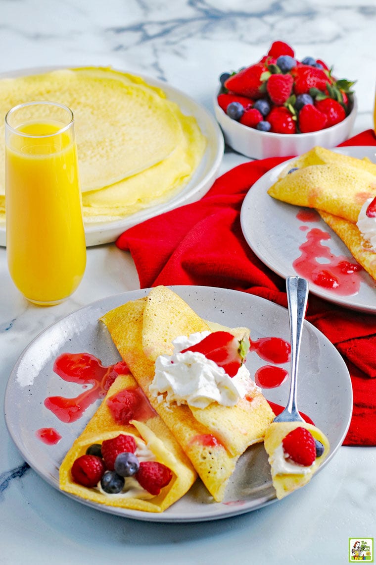 A berry and cream filled crepes on a plate with plates of gluten free crepes, a bowl of berries and a glass of orange juice on a red cloth napkin.