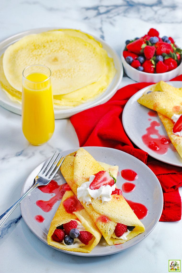 A tabletop with plates of gluten free crepes with berries, whipped cream, a glass of orange juice, and a bowl of raspberries and blueberries.