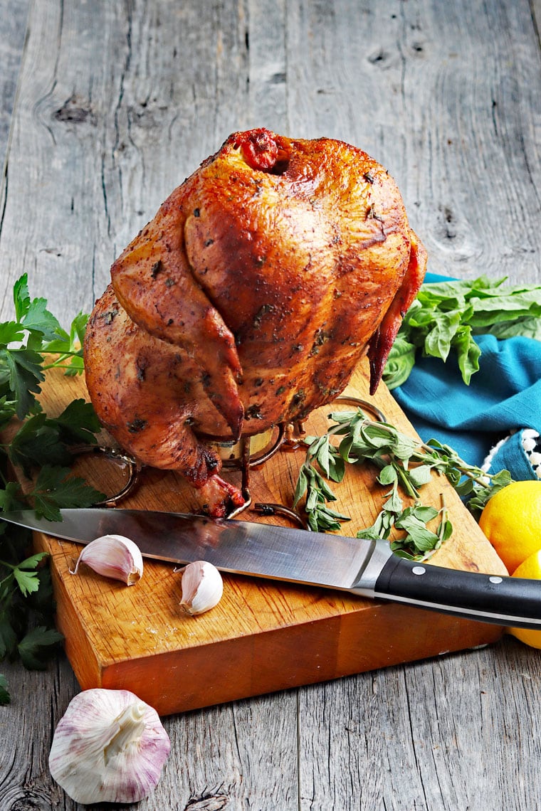 Smoked beer can chicken on a roaster rack placed on a cutting board with a knife with garlic cloves and herbs.