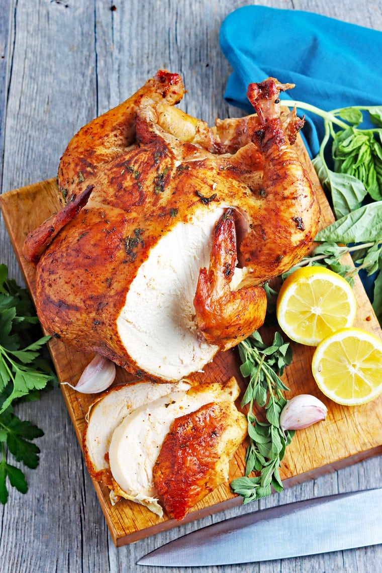 Sliced roasted chicken on a wooden cutting board with lemons, garlic and herbs with a blue cloth napkin.