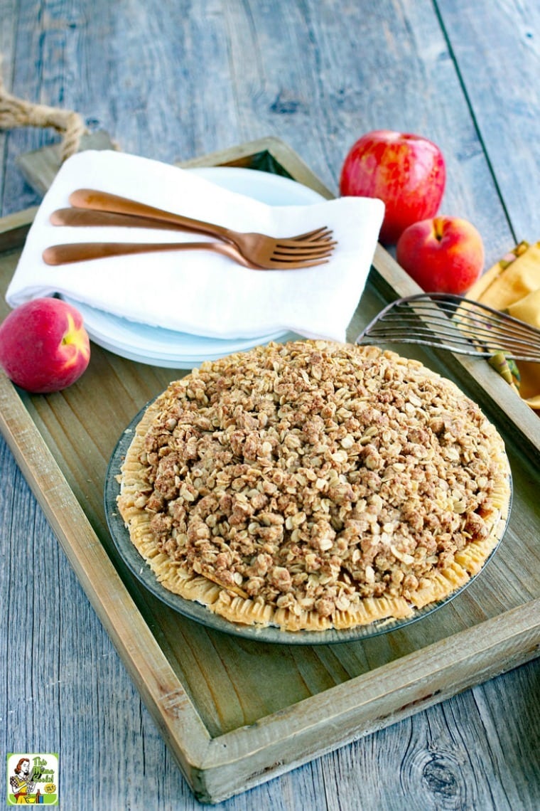 Pie with crumb topping on a wooden tray with white plates, white napkins, copper forks, red apples, yellow tea towels, and a vintage spatula.