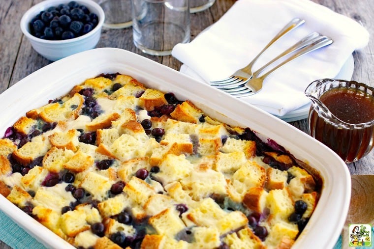 Blueberry French Toast Casserole in a large dish with maple syrup, bowl of blueberries, plates, napkins, glasses, and forks.