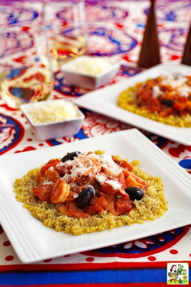 Bowls of shrimp puttanesca on a colorful tablecloth, glasses of white wine, and small bowls of shredded cheese.