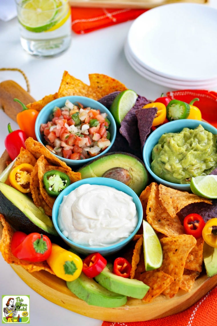 A platter with a small blue bowl of vegan sour cream with slices of limes, tortilla chips, mini sweet peppers, avocados, blue bowls of salsa and guacamole, and white plates.