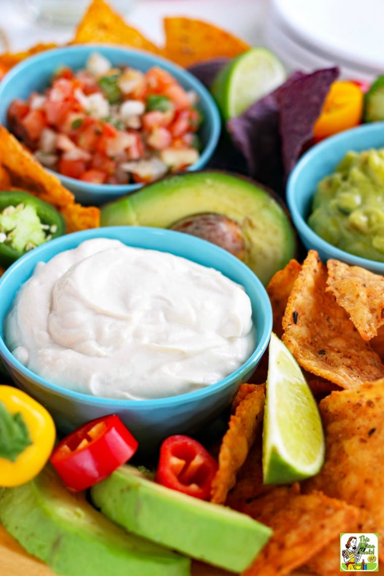 Closeup of blue bowls of creamy dairy-free sour cream, salsa, and guacamole with slices of lime, tortilla chips, sweet peppers, and avocados.