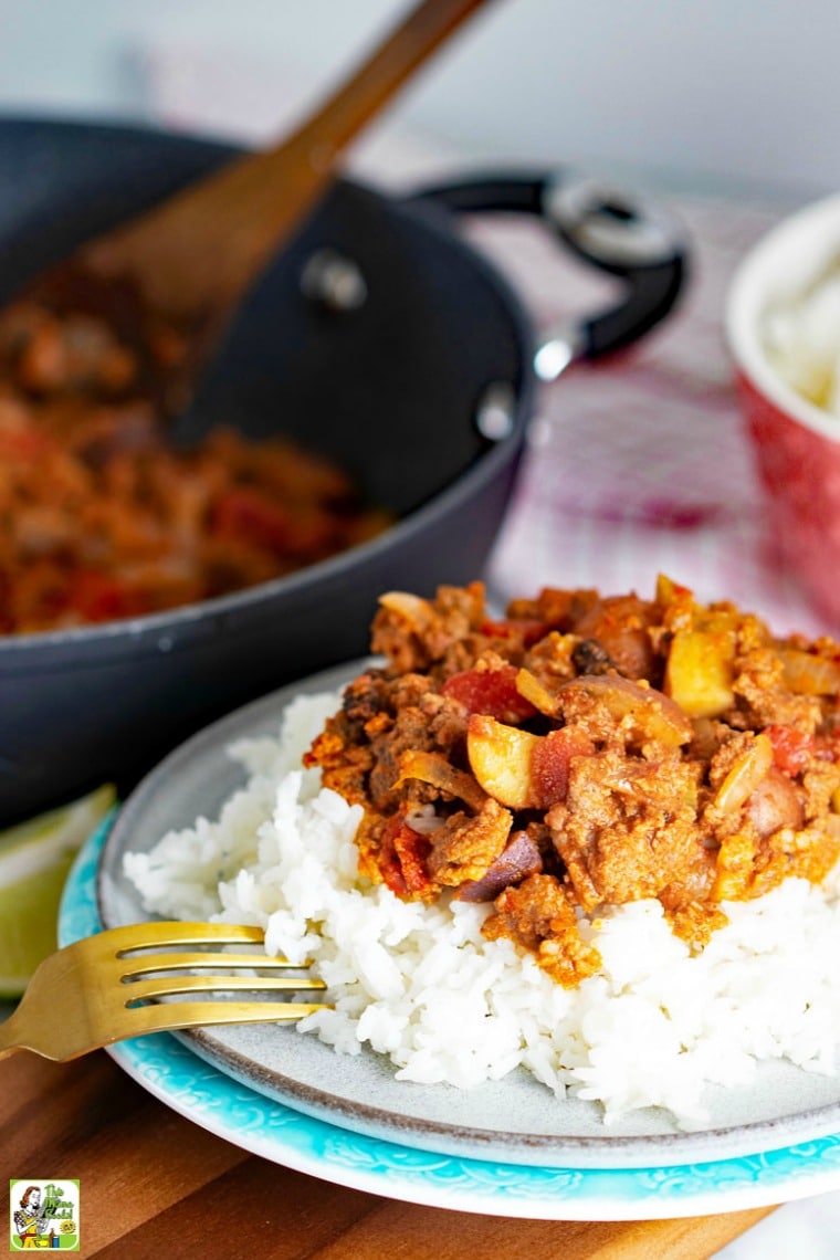 Plate of rice and Mexican Picadillo with fork and saute pan and wooden spoon and pink bowl in the background.