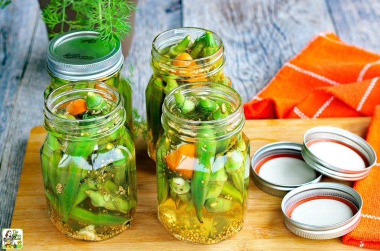 Jars of Refrigerator Pickled Okra with lids on a wooden cutting board with orange cloth napkins.