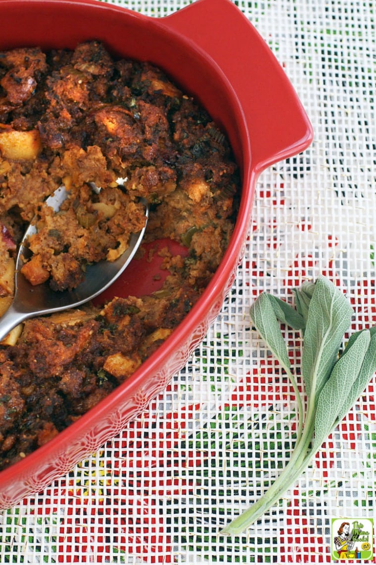 A red casserole dish of Thanksgiving stuffing with serving spoon and a sprig of sage on a festive holiday tablecloth.
