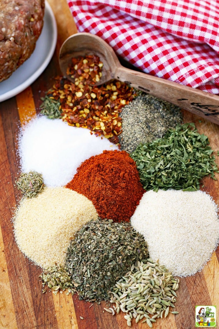 Small mounds of spices and seasonings, a measuring spoon, a checked napkin, and the edge of a bowl with ground meat, sitting on a wooden cutting board.