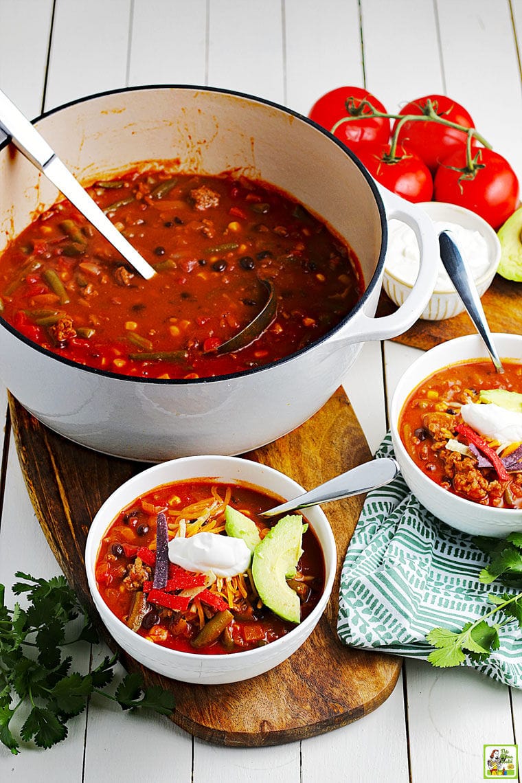 A large Dutch oven of taco soup with cooking spoon and two white bowls of soup with spoons on a wooden cutting board with a napkin with tomatoes in the background.