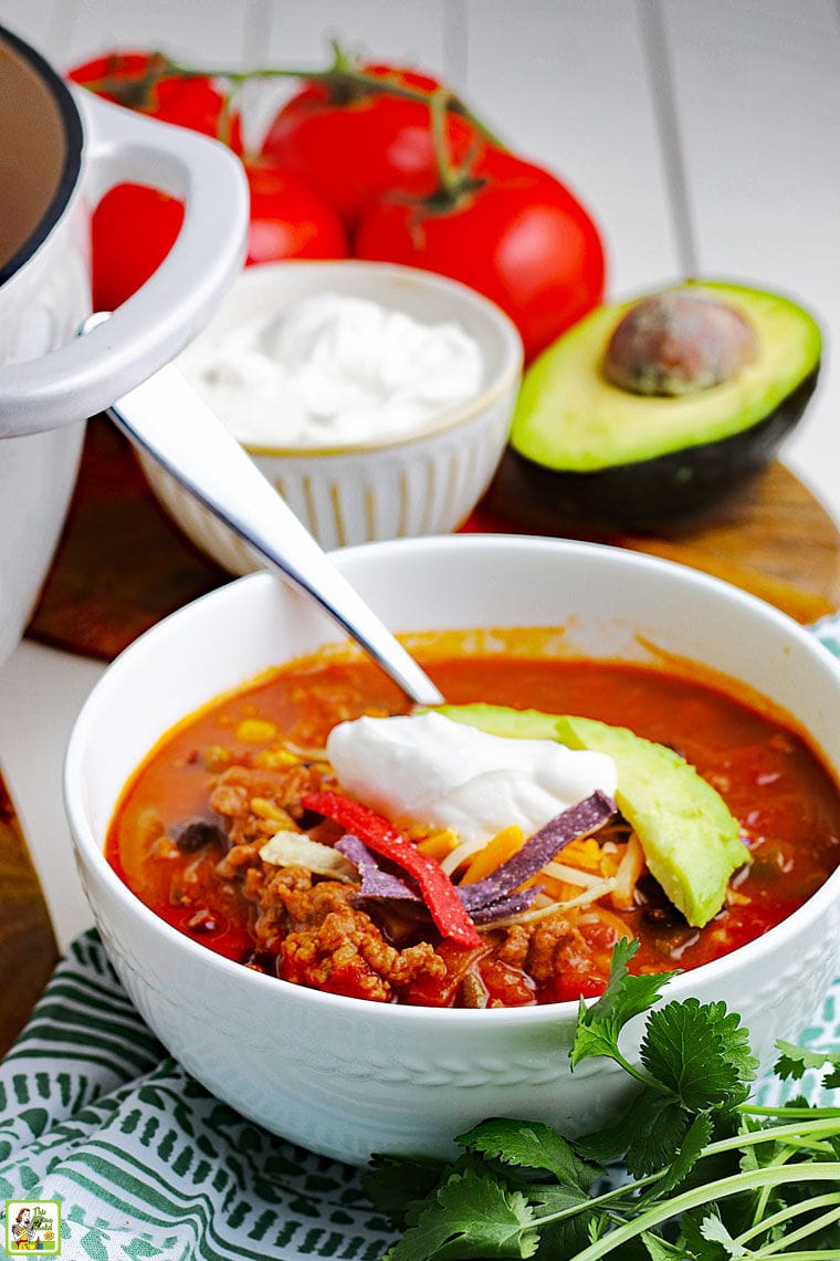 Closeup of a bowl of taco soup with spoon, dollop of sour cream, slice of avocado, and pieces of tortilla chips with a bowl of sour cream, avocado and tomatoes in the background.