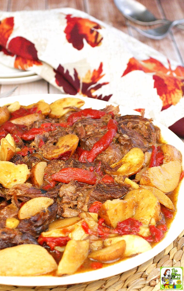 A plate of pot roast with red peppers and potato wedges with a floral napkin and white plates in the background.