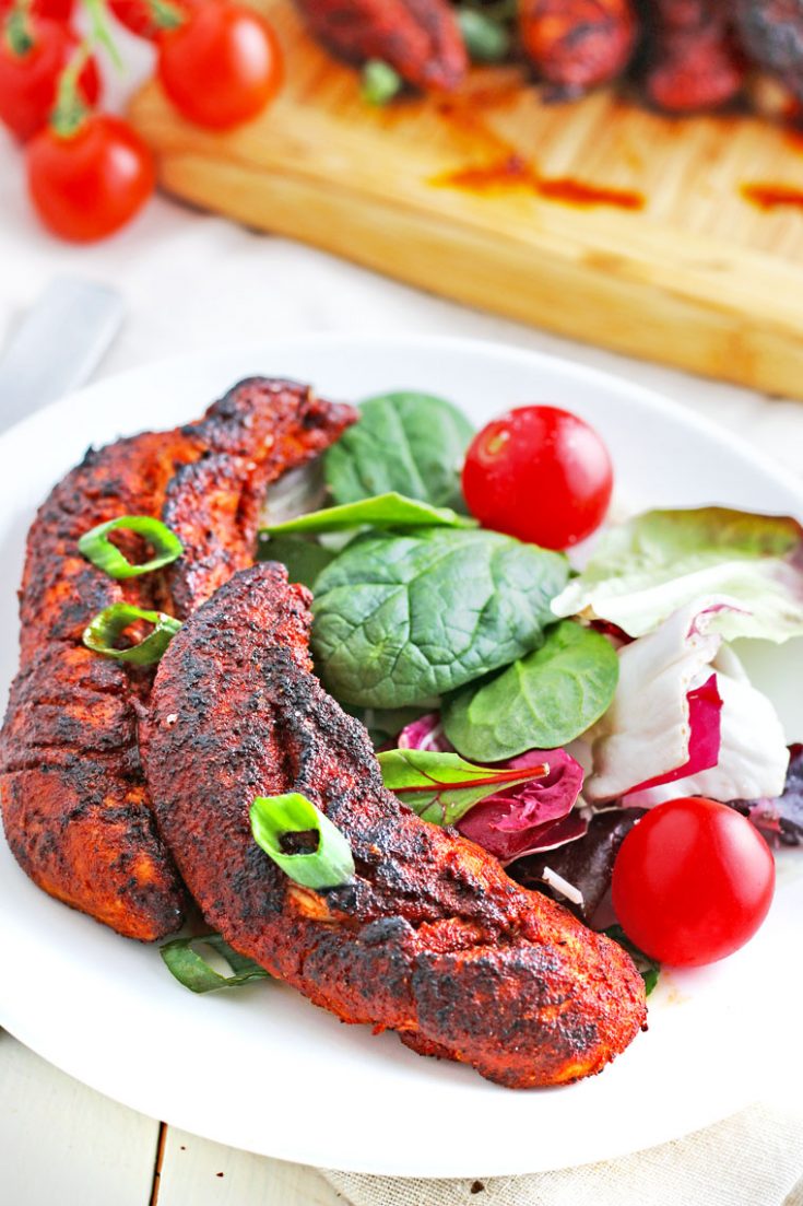 A white plate with two blackened chicken tenders, lettuce, and tomatoes.