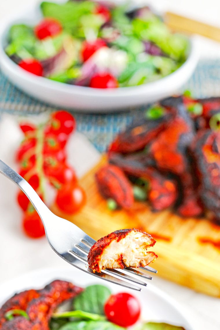 Closeup of a fork with a piece of cooked chicken with a bowl of salad, tomatoes, and blackened chicken tenders on a wooden board in the background.