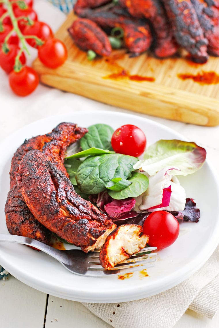 A plate of chicken tenders with a side salad and fork with tomatoes and chicken on a wooden cutting board in the background.