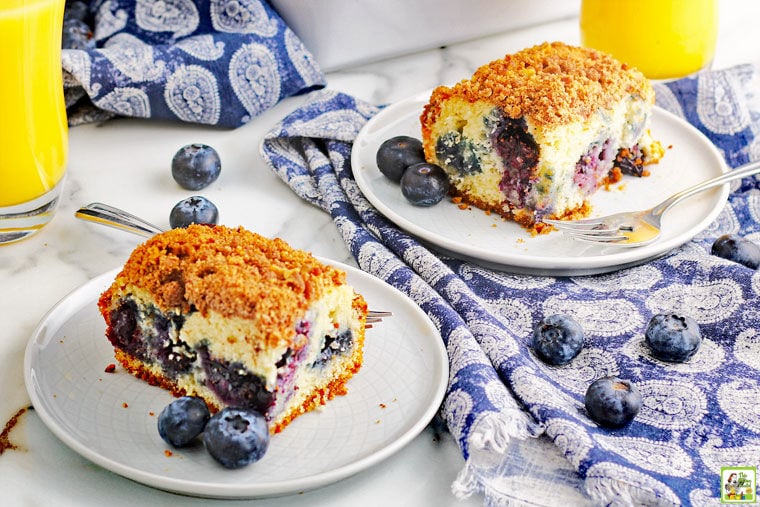 Two white plates of blueberry muffin cake with forks, blue napkins, glasses of orange juice, and blueberries.