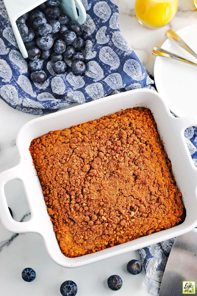 Overhead shot of a baking dish of blueberry muffin cake with blue napkin, glass of orange juice, white plates, forks, cake server, and blueberries.