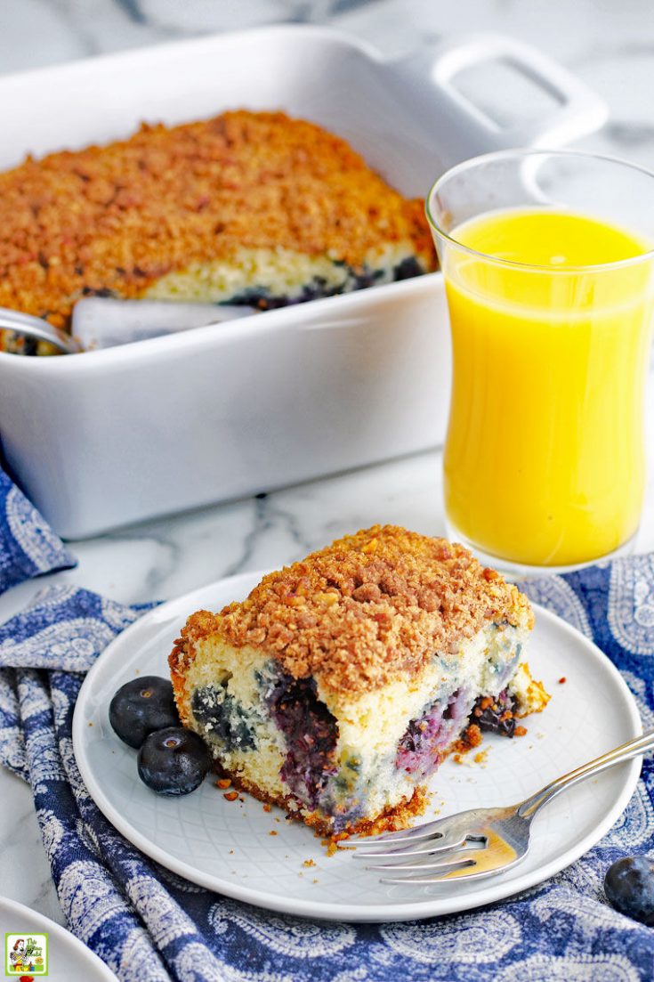 A piece of blueberry muffin coffee cake on a white plate with fork, a baking dish of coffee cake, and a glass of orange juice on a blue and white napkin.