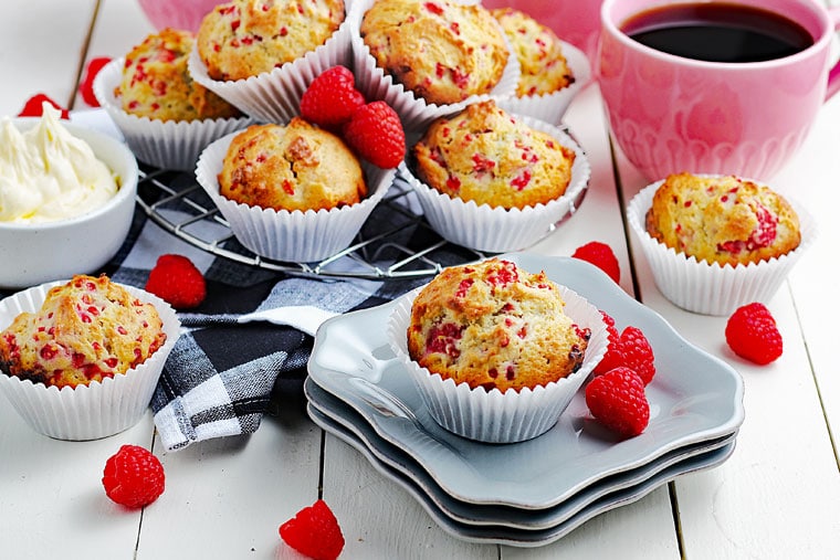 A tabletop of raspberry yogurt muffins with a black and white napkin, fresh raspberries, white plates, a bowl of butter, and pink mugs of black coffee.
