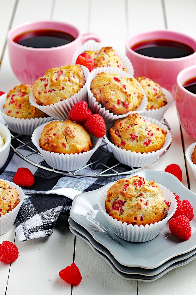 A tabletop with muffins in white cupcake liners on white plates and on black and white napkins with red raspberries and pink mugs of black coffee.