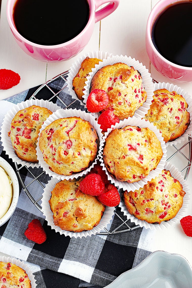 Overhead shot of a bunch of raspberry muffins in white paper liners on a wire rack on a black and white napkin with mugs of coffee.