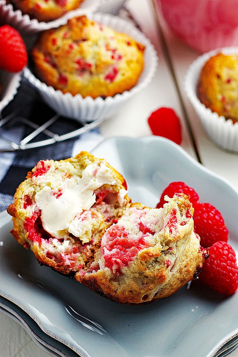 Closeup of a raspberry muffin torn in half filled with a dollop of butter on a gray plate with fresh raspberries.