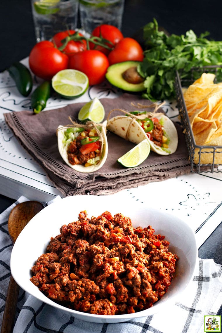 A white dish of spicy Crockpot Taco Meat with tacos, limes, tortilla chips, tomatoes, chilis, and cilantro.