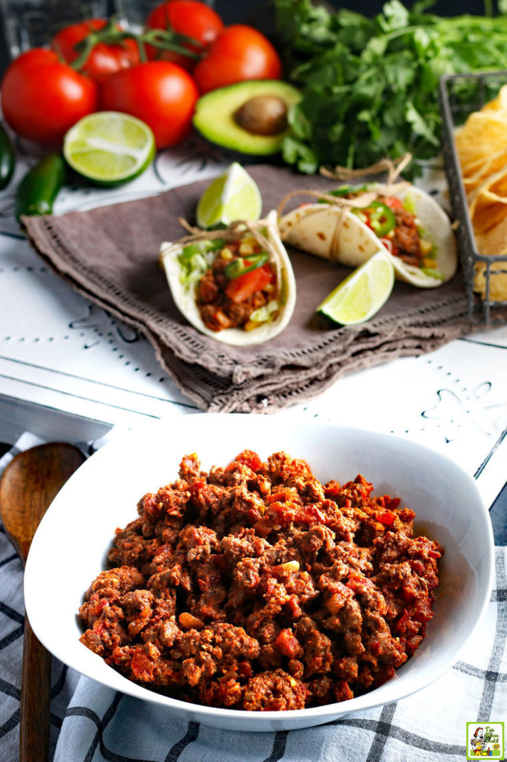 A white bowl of Crockpot Taco Meat with slow cooker tacos, limes, tomatoes, and avocados