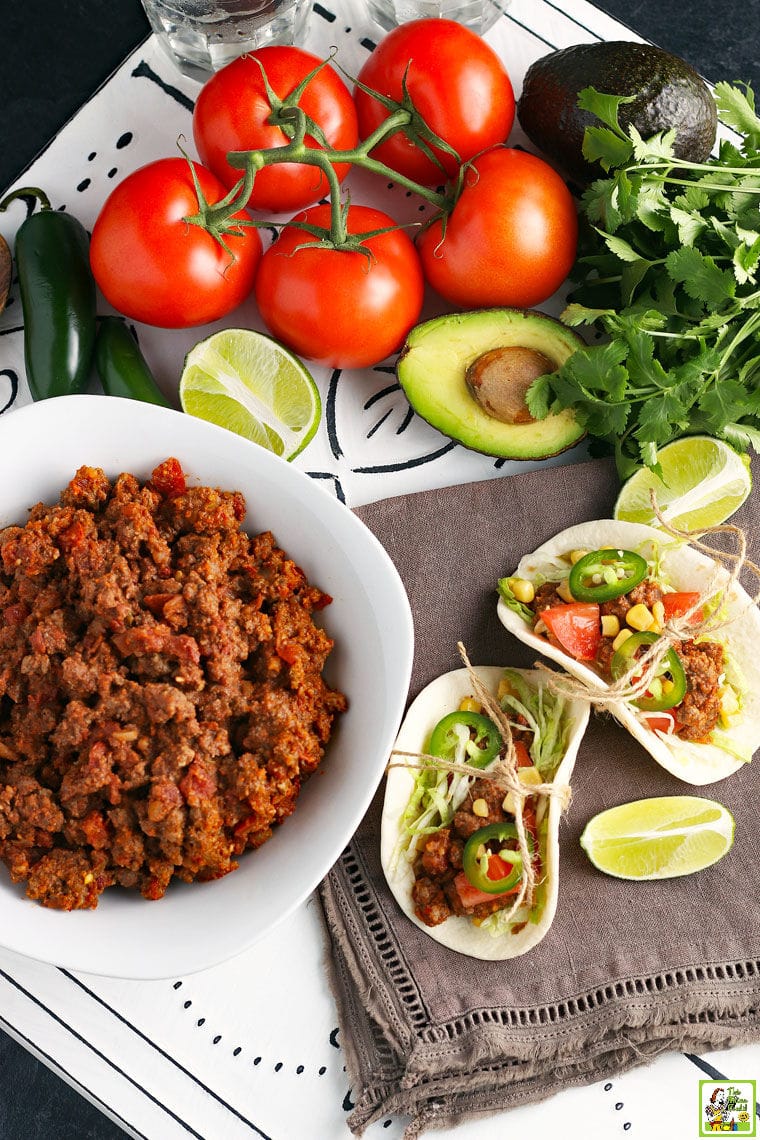 A bowl of cooked Crockpot Taco Meat with crock pot tacos, limes, tomatoes, and avocados.