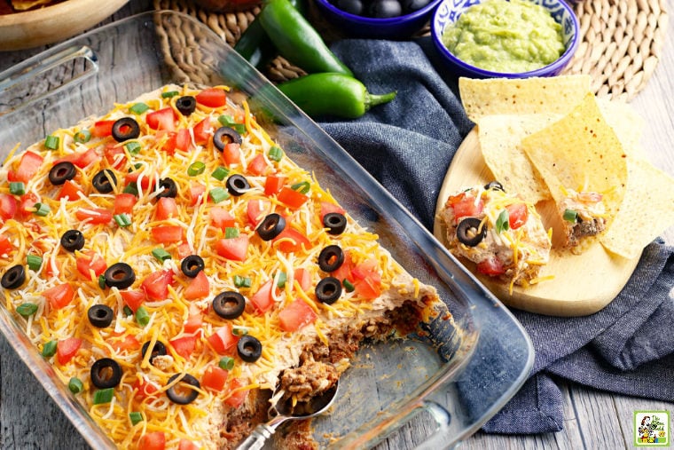 Layered taco dip in a casserole dish with serving spoon, chili peppers, bowls of guacamole, tortilla chips, and napkins.