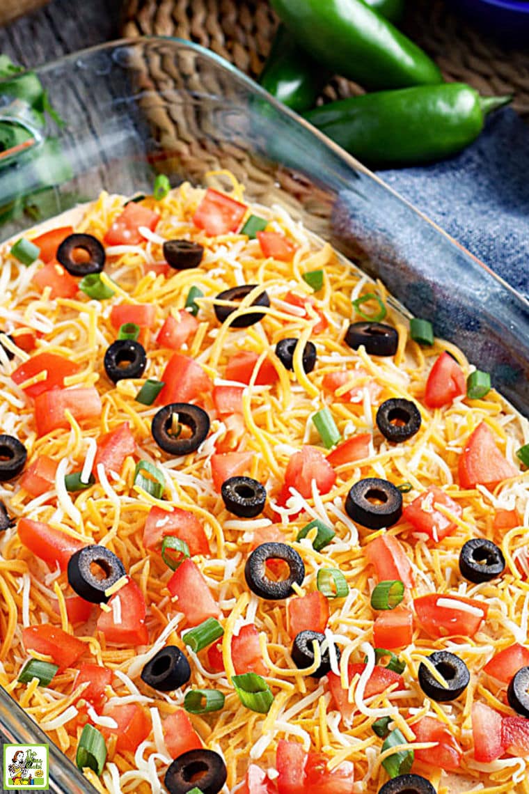 A glass casserole dish of taco dip with cheese, olives, tomatoes, and green onions.