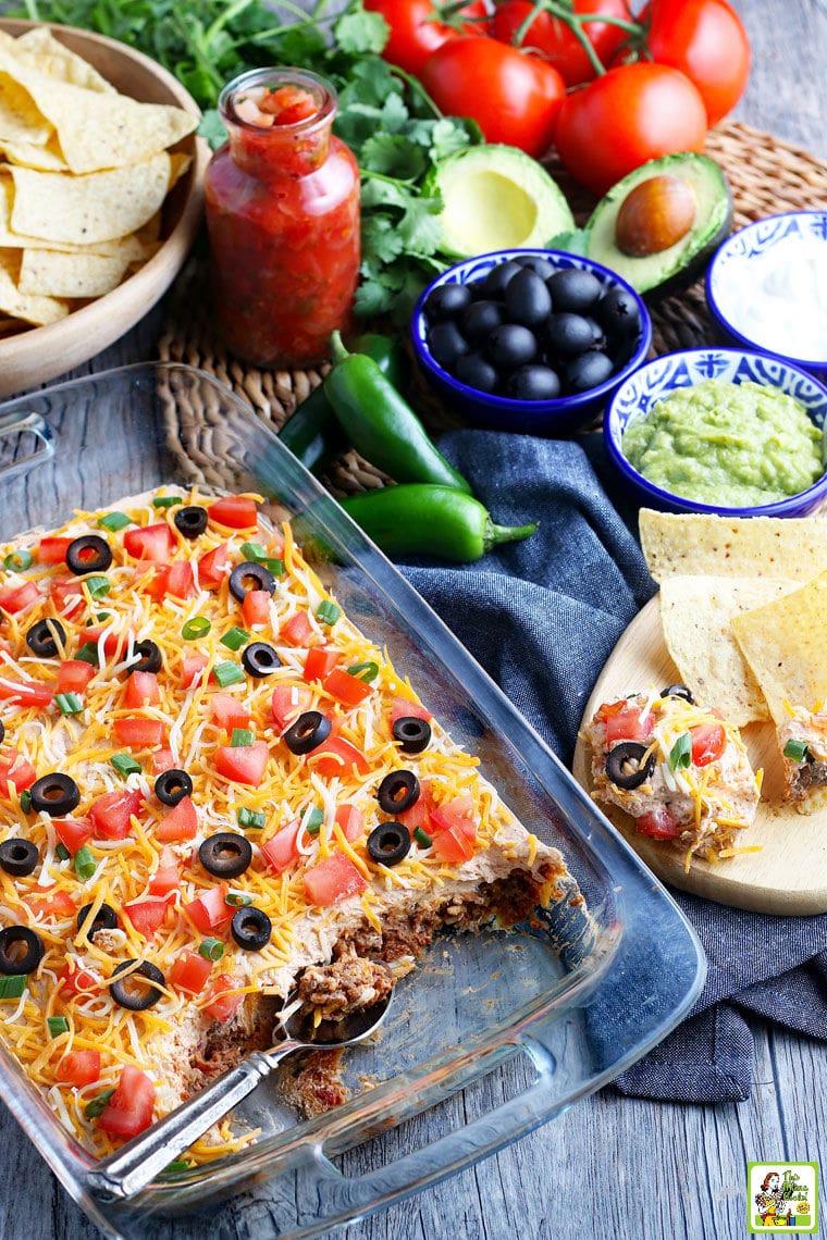 A casserole dish of dip with cheese, olives, tomatoes, and green onions layered with sour cream, refried beans and ground beef.