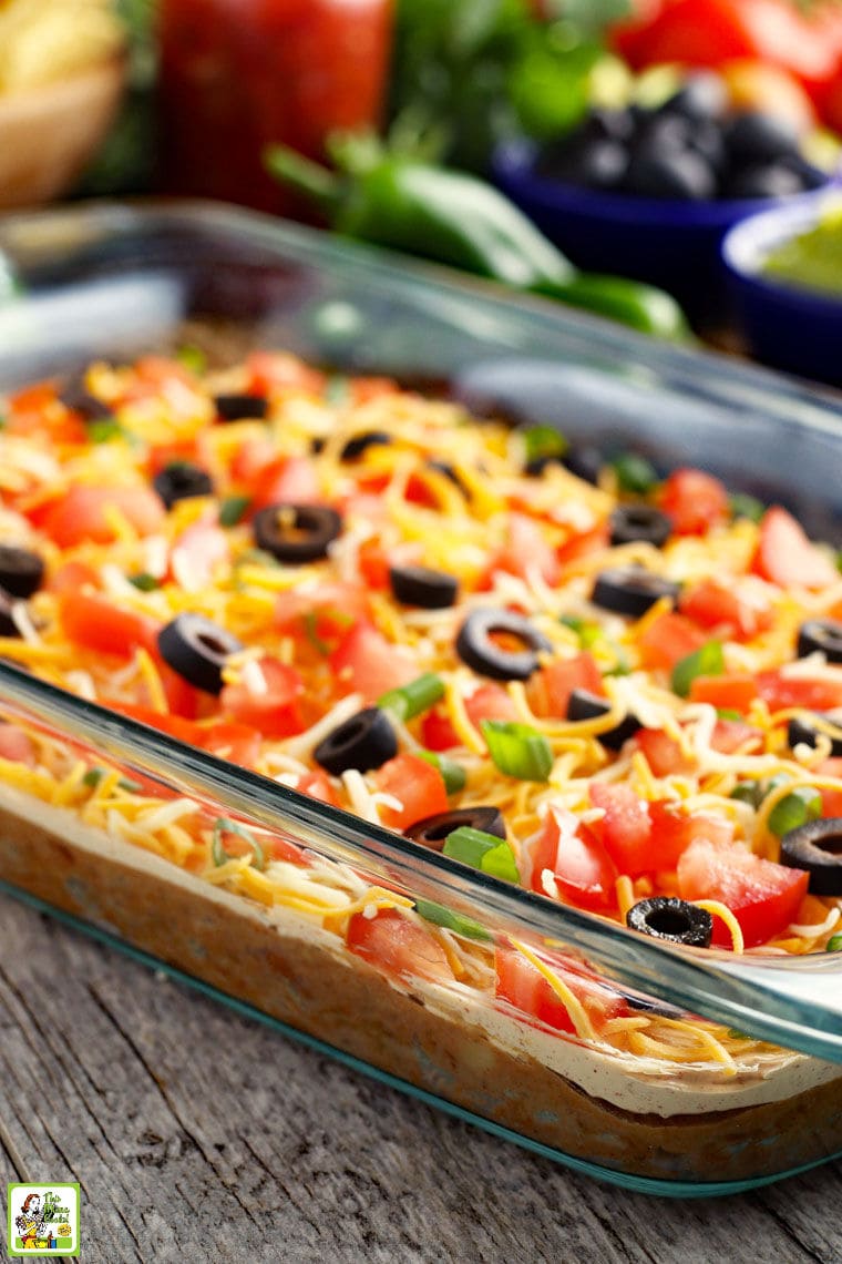 A glass casserole dish of Mexican taco dip with layers of cheese, olives, tomatoes, green onions, sour cream, refried beans, and ground beef.