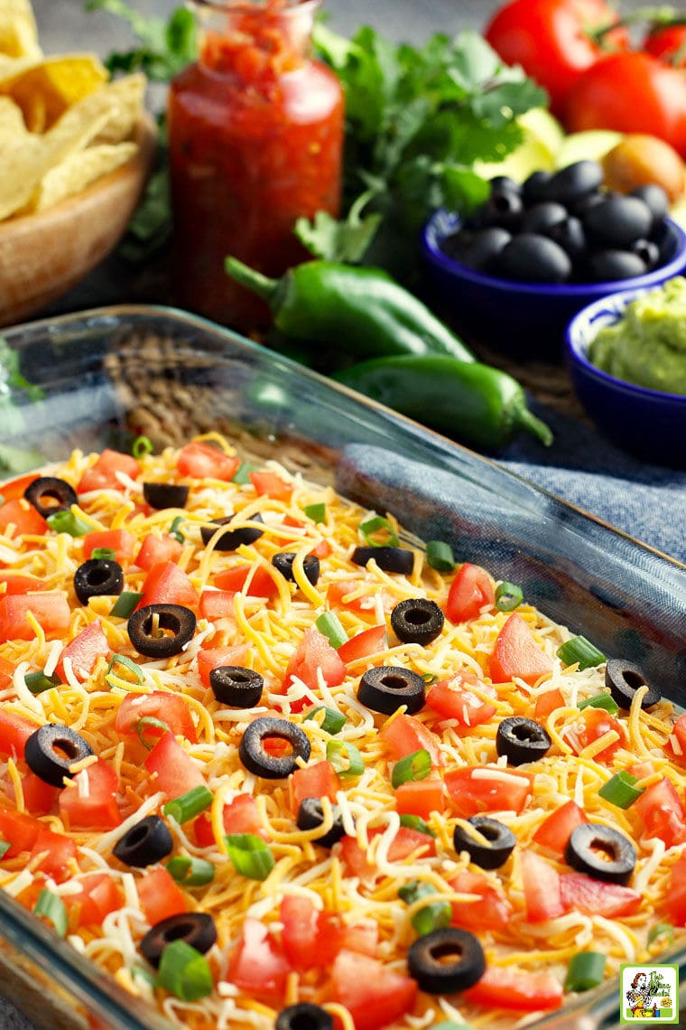 A casserole dish of cheese, olives, tomatoes, and green onions. With olives, chiles, and tomatoes in the background.