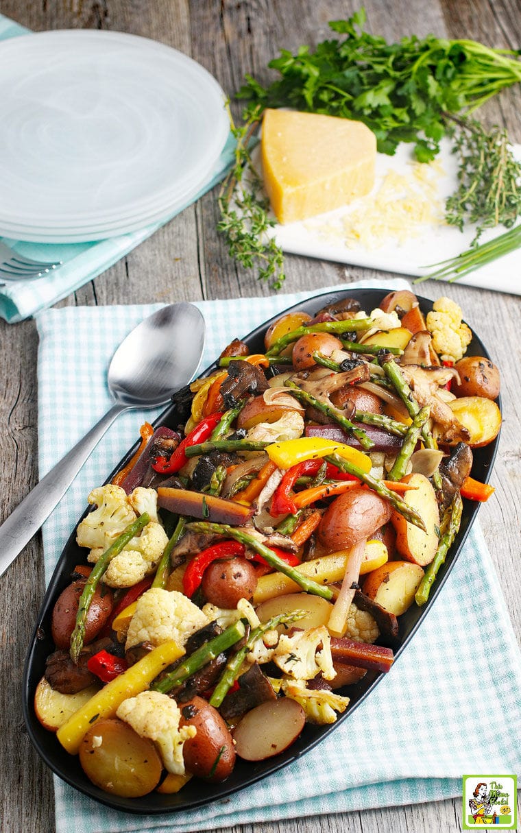 A platter of oven roasted veggies like potatoes, asparagus, cauliflower, and bell peppers placed on a napkin with a spoon, with herbs and cheese in the background.