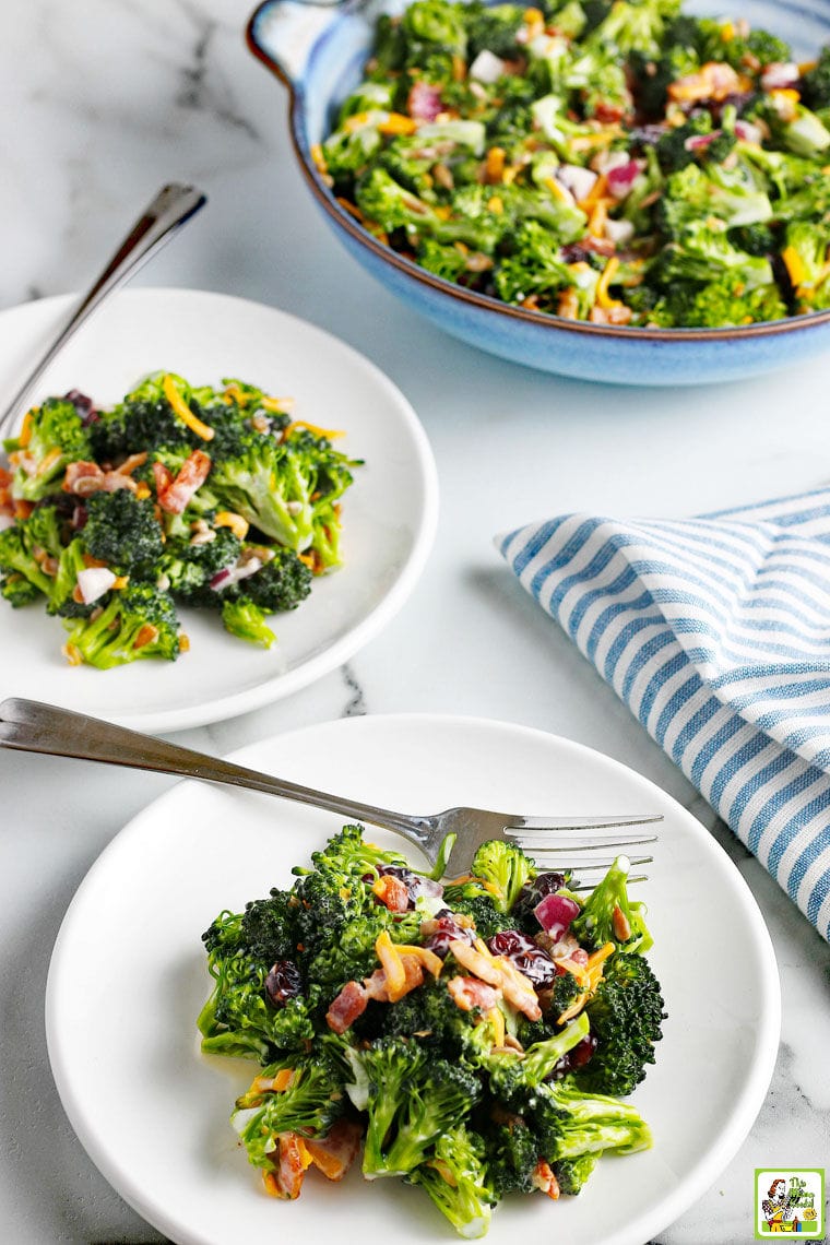 Broccoli Salad with cheese, onion and cranberries on white plates with forks and blue and white napkins. With a blue salad bowl in the background.