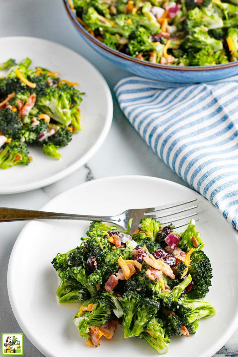 Broccoli Salad with cheese, onion and cranberries on white plates with forks and blue and white napkins. With a blue salad bowl in the background.