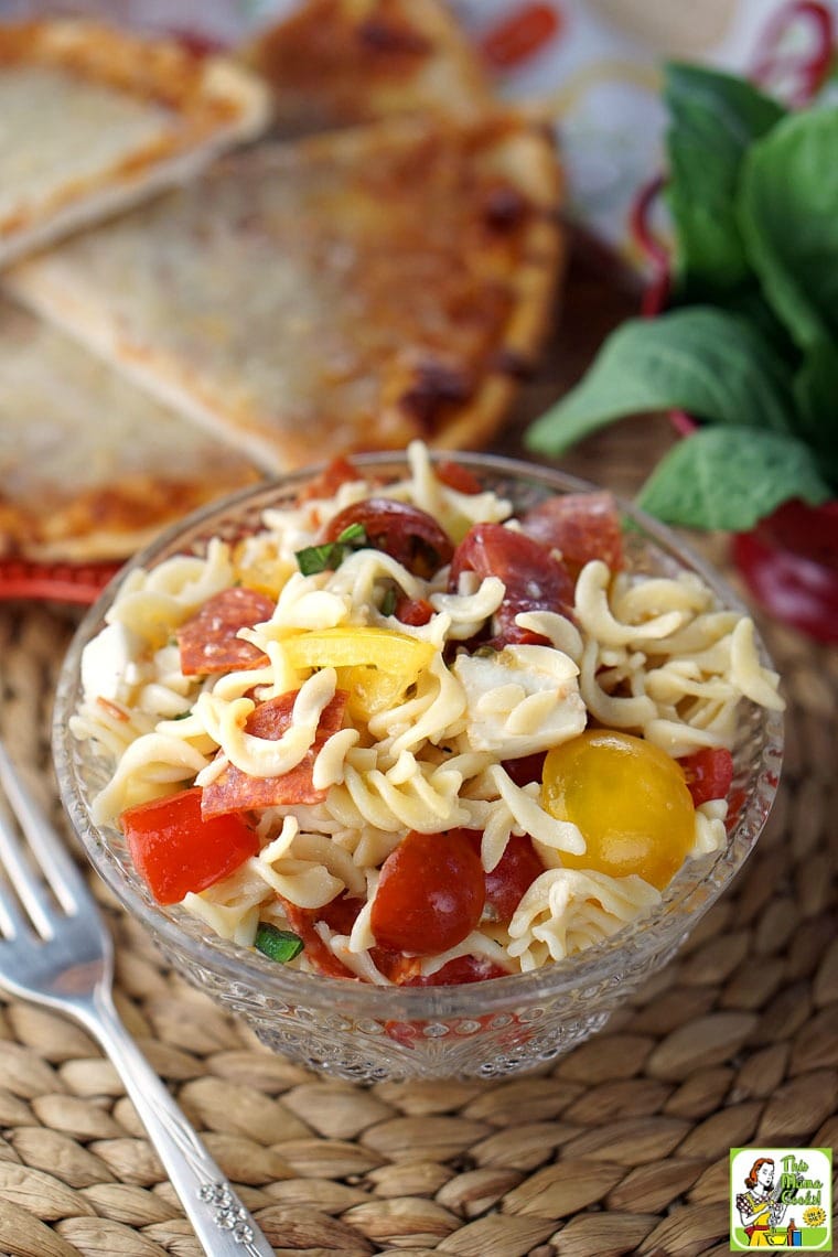 A glass bowl of Caprese Pasta Salad with fork with slices of pizza in the background.