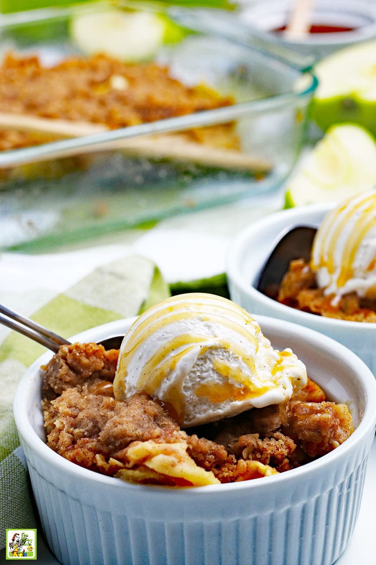 A white bowl of warm apple crisp with a scoop of vanilla ice cream drizzled with caramel sauce and a spoon.