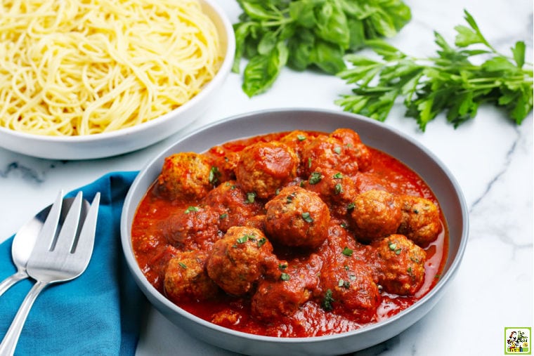 A large bowl of Instant Pot Meatballs in sauce, a large bowl of spaghetti, fresh sprigs of parsley and basil, serving utensils, and a blue cloth napkin.